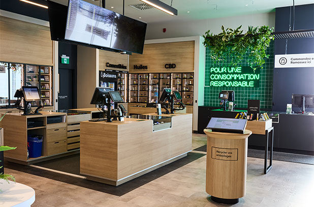 Intérieur moderne d'une succursale de la SQDC, avec des comptoirs de service en bois clair, des écrans de commande, une section de ramassage, et un mur vert illuminé portant le message 'Pour une consommation responsable'.